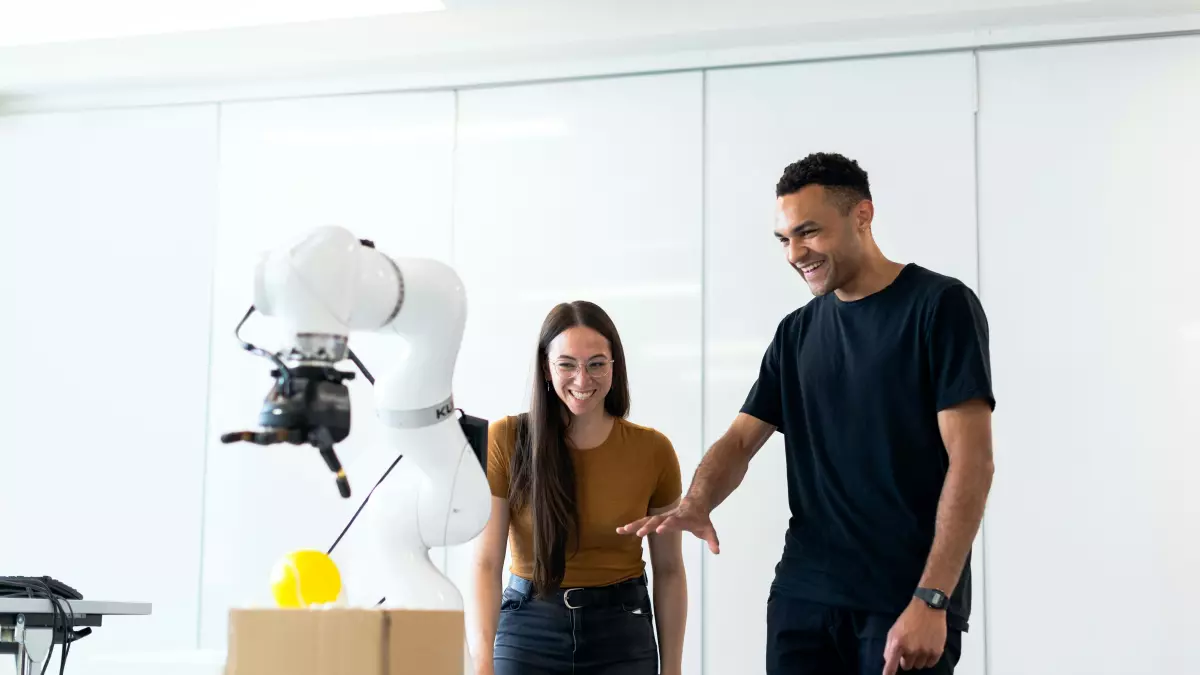 Two people look intently at a robotic arm moving a yellow object to a cardboard box.