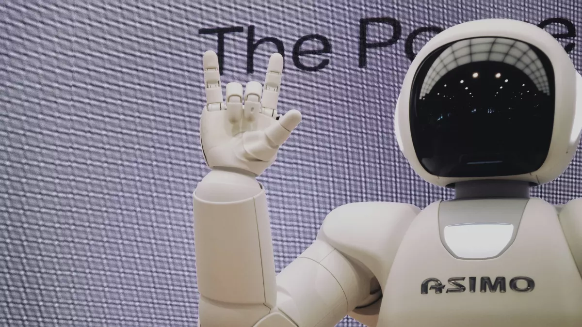 A humanoid robot with a white body and a black head, its right arm raised in a gesture, the background is blurred.