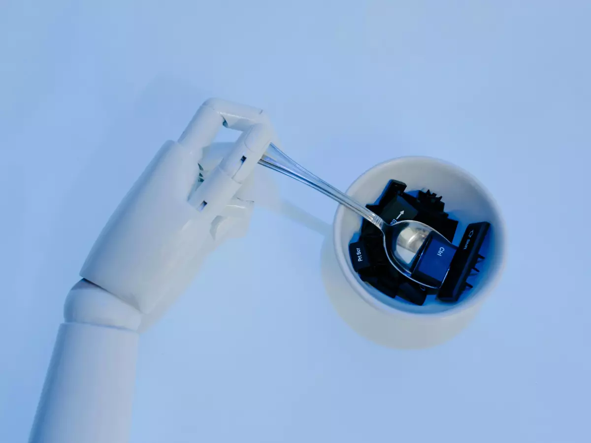 Close-up of a robotic hand, with delicate fingers, holding a small transparent sphere with blue and black internal components. The hand is white against a light blue background, emphasizing the mechanical precision.