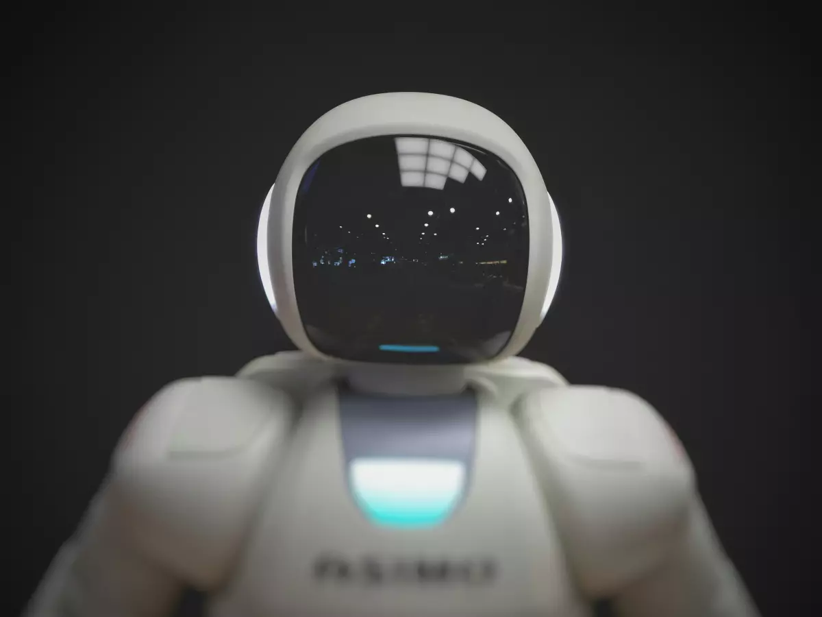 A close-up of a white humanoid robot's face with a neutral expression. The robot has a smooth, plastic face with large black eyes and a small mouth. The background is black, with a faint reflection of lights.