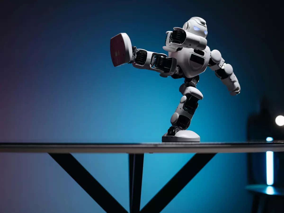 A white robot with black accents is shown kicking its leg while standing on a table. The background is blue, and the table is made of glass. The robot's eyes are blue, and it has a white head. It is wearing a white shirt and black pants. It is holding a white ball in its left hand. It is standing on a white surface, and the floor is also white. The image is taken from a high angle.