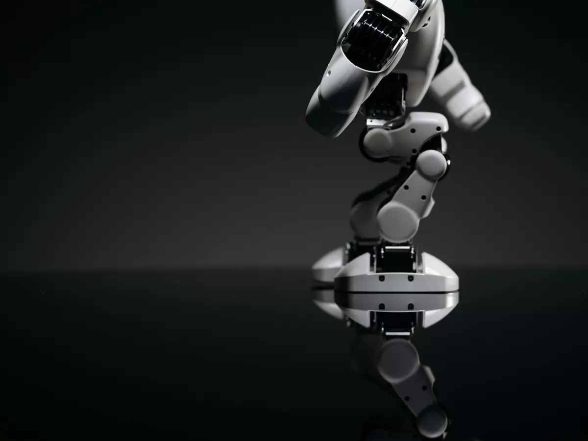 Close-up shot of a white robot's legs and body, showcasing its joints and mechanisms. The robot is posed dynamically, suggesting movement and flexibility. The background is black with a reflective surface creating a dramatic effect.