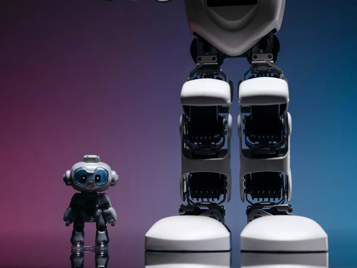 A white humanoid robot with black accents stands tall, its legs and feet prominently displayed.  A smaller humanoid robot, facing the viewer, stands at the base of the larger robot's legs. The backdrop features a gradient of deep blue to pink. 