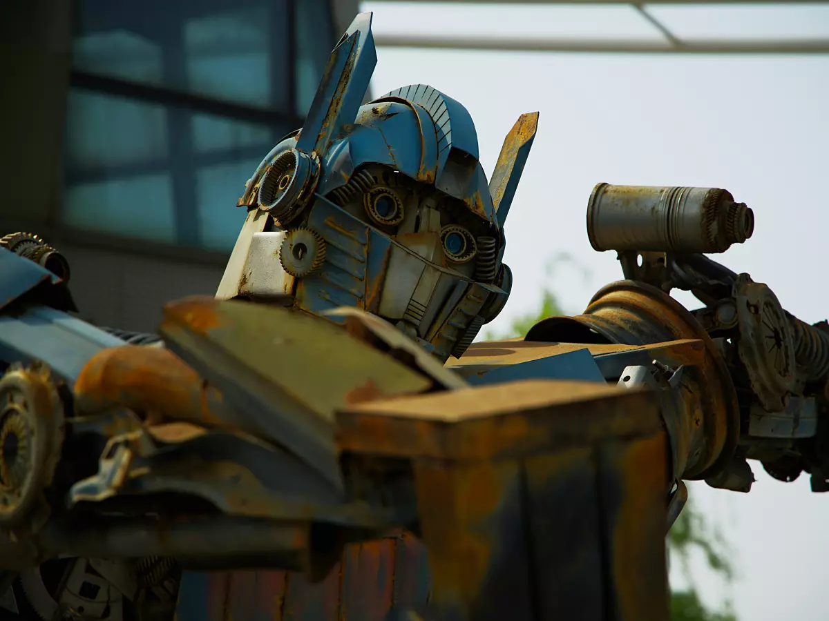 Close-up of a large humanoid robot made of scrap metal, resembling a Transformer, with a blue, yellow, and rust color scheme. The robot is positioned against a windowed building background.