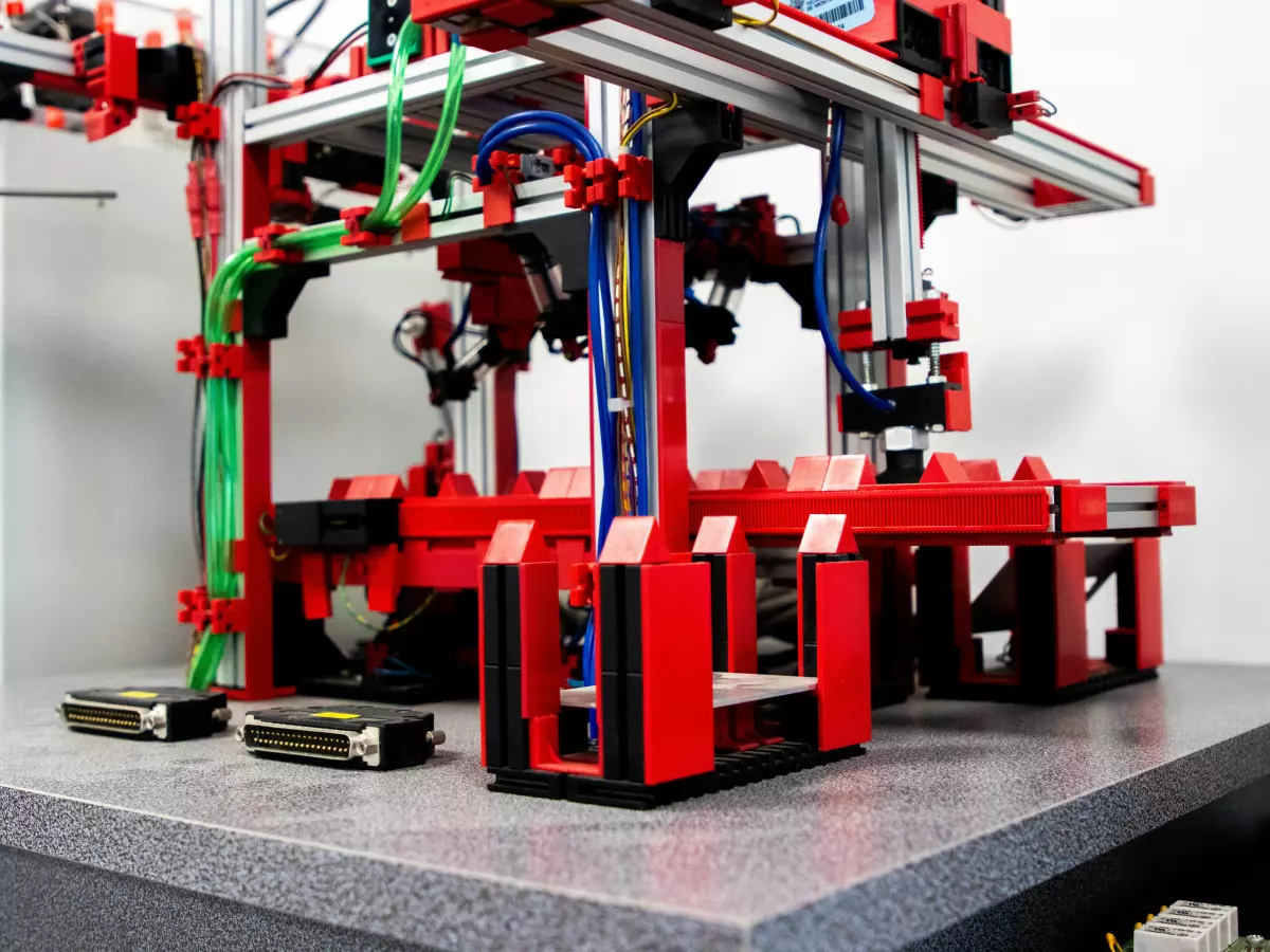 A robotic arm, comprised of red metal segments and cables, moving on a platform. The metal parts of the robot are partially covered by red, blue, and green cables.