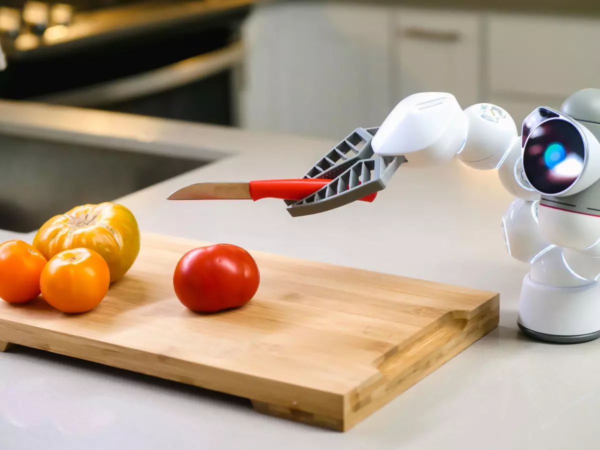 A white humanoid robot with a robotic arm holding a knife on a wooden cutting board with a tomato and a few oranges nearby.