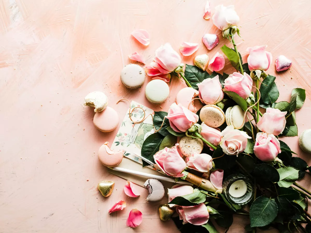 A bouquet of pink roses, some macaron cookies, and golden and pink chocolates lie on a light pink background.