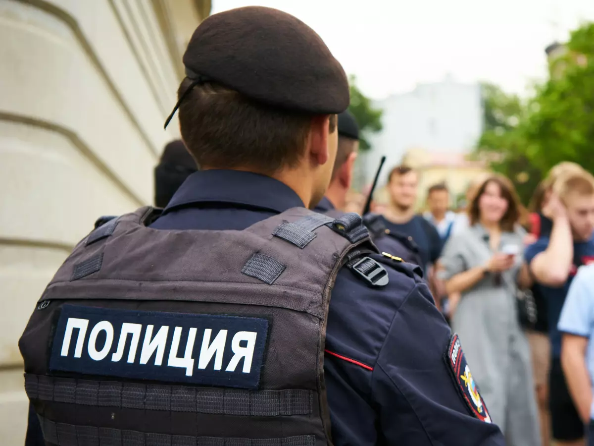 A police officer in full gear with a 'POLICE' inscription on his vest. The officer is facing away from the camera and looking at a crowd of people in the background.