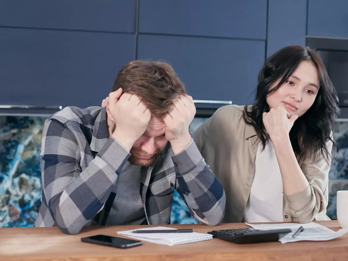 A man sitting at a table with his head in his hands, a woman sitting next to him looking concerned. The man is wearing a plaid shirt and the woman is wearing a black shirt.