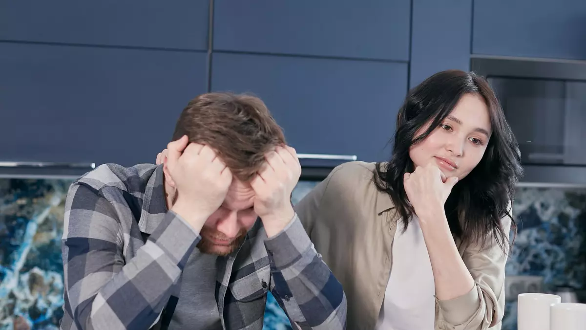 A man sitting at a table with his head in his hands, a woman sitting next to him looking concerned. The man is wearing a plaid shirt and the woman is wearing a black shirt.