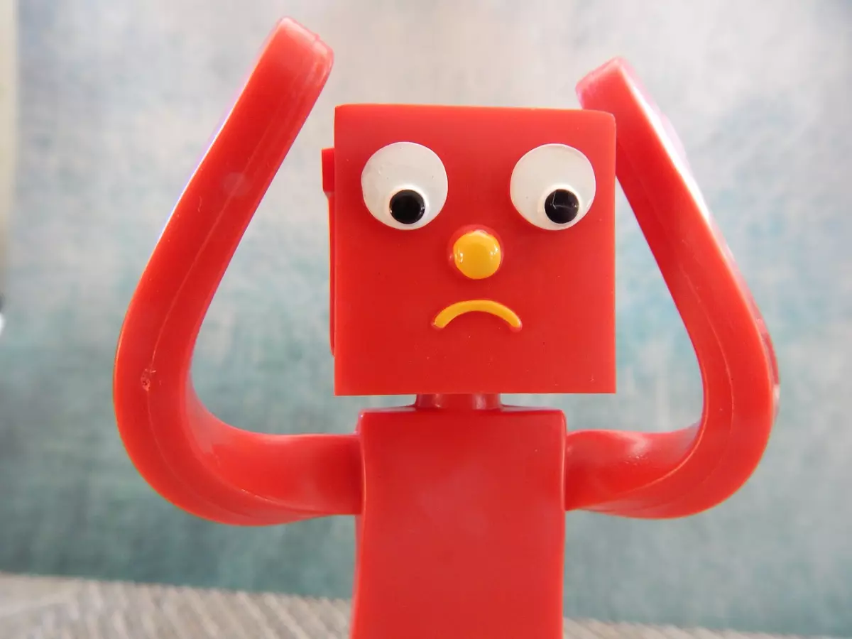 A red plastic toy robot with big eyes and a yellow nose looks frustrated. It has its hands raised as if it is pulling its hair out.