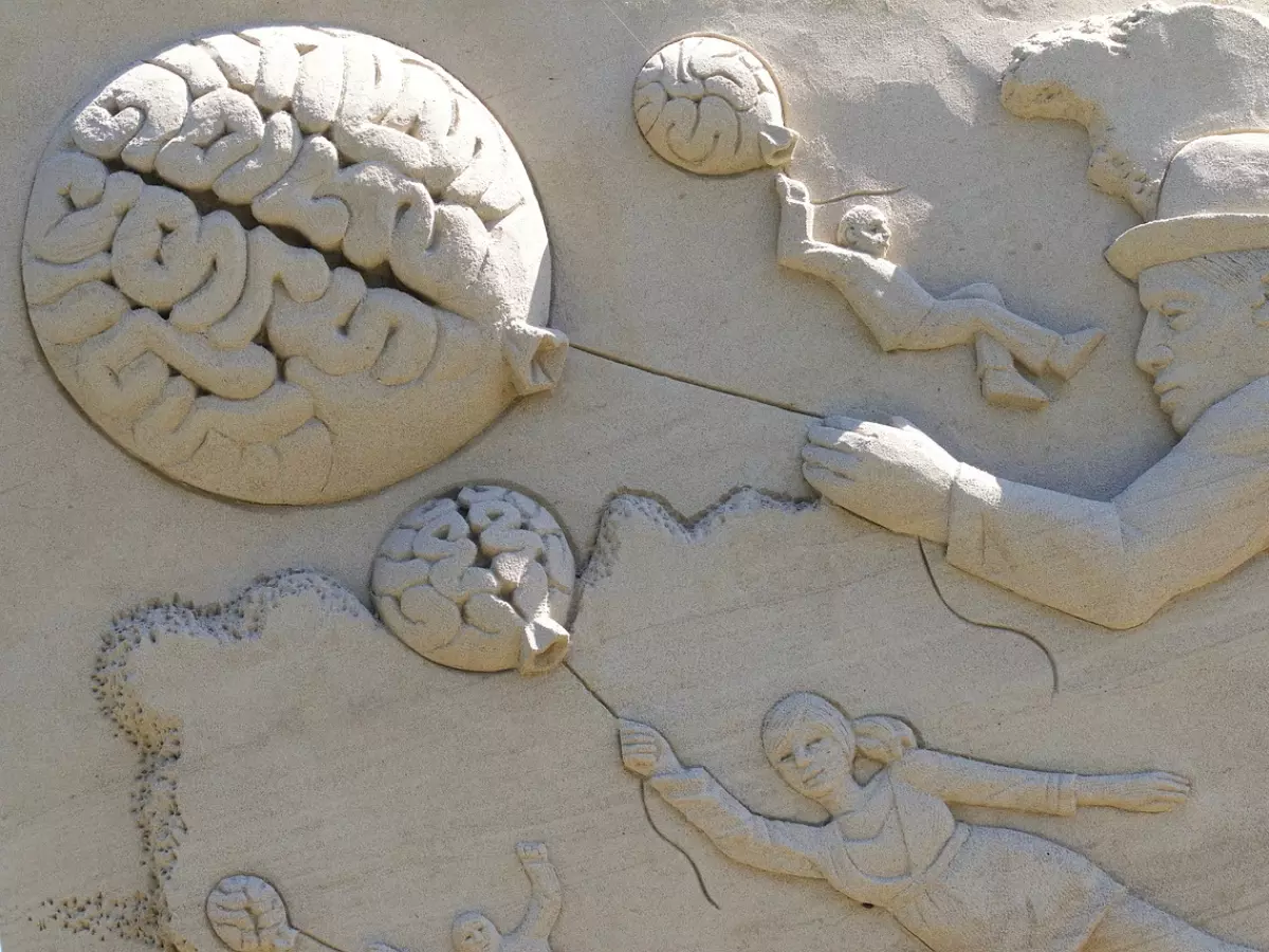 A sculpture of a man holding a balloon shaped like a brain. There are other sculptures of people and brains in the background. The sculpture is made of sand.