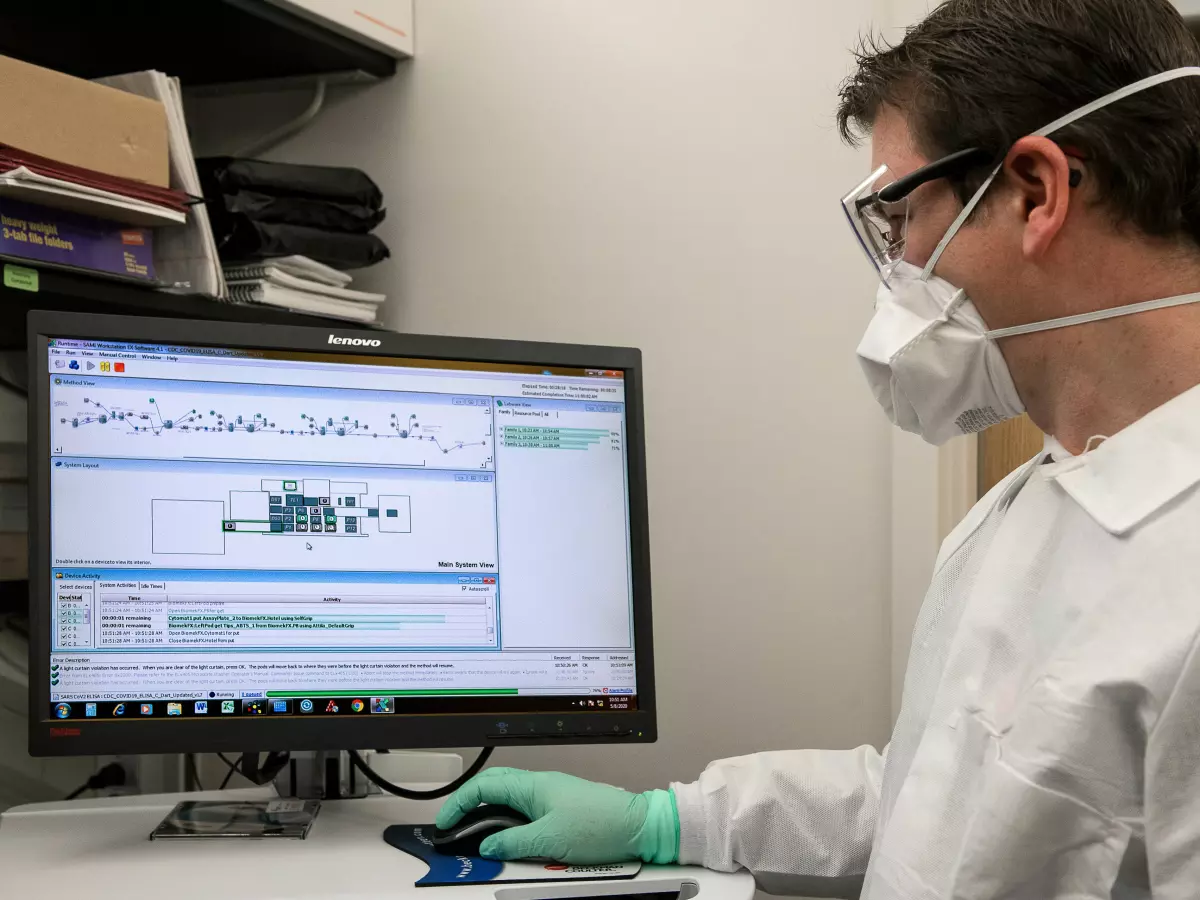 A scientist in a lab coat and protective gear is working on a computer, with a data screen in front of him. 