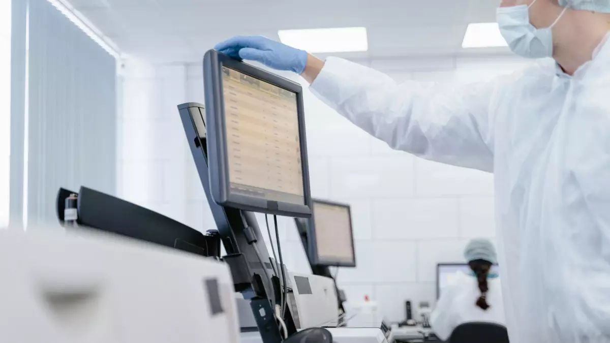 A scientist in a lab coat adjusts a computer monitor displaying data; multiple monitors and lab equipment in the background.