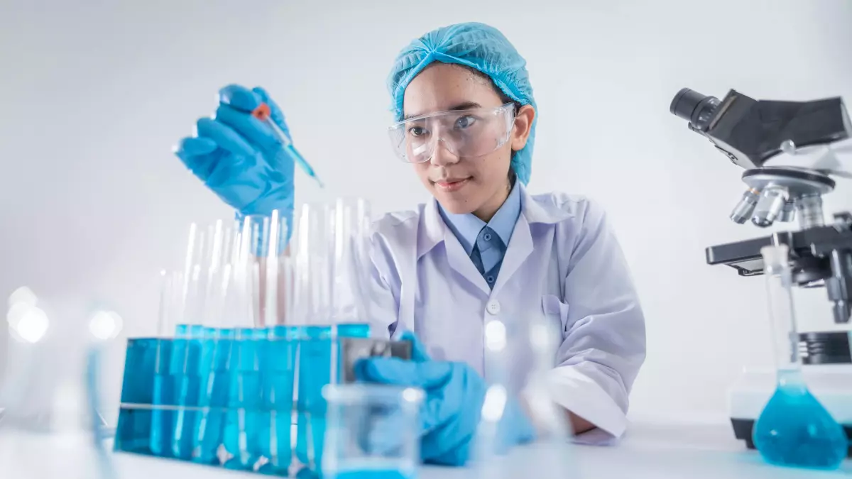 A woman in a lab coat, gloves, and a hairnet is using a pipette to transfer a liquid from one test tube to another,  in a laboratory setting. There are other test tubes, beakers, and a microscope in the background.