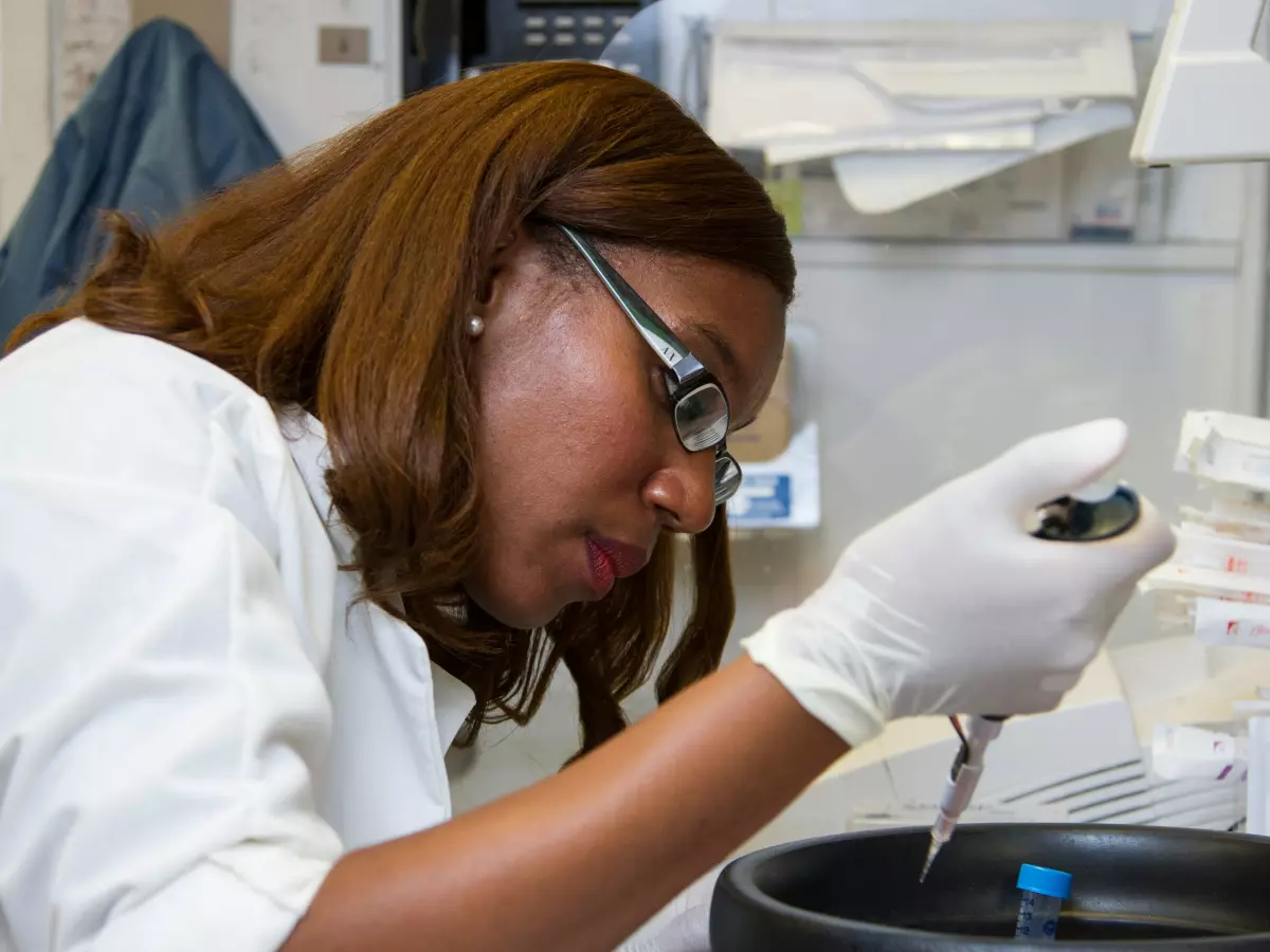 A woman in a lab coat is looking through a microscope, with various beakers and lab equipment in the background.