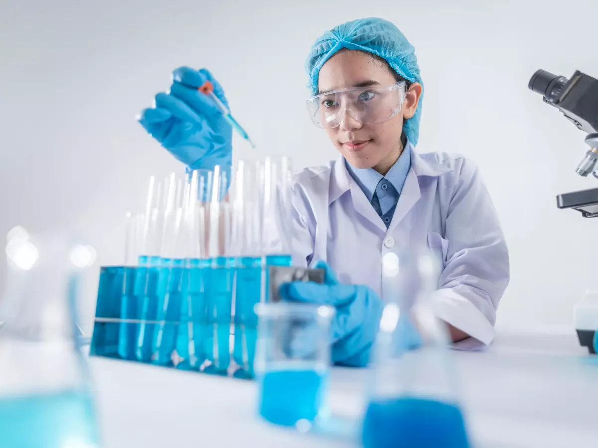 A woman in a lab coat, gloves, and a hairnet is using a pipette to transfer a liquid from one test tube to another,  in a laboratory setting. There are other test tubes, beakers, and a microscope in the background.