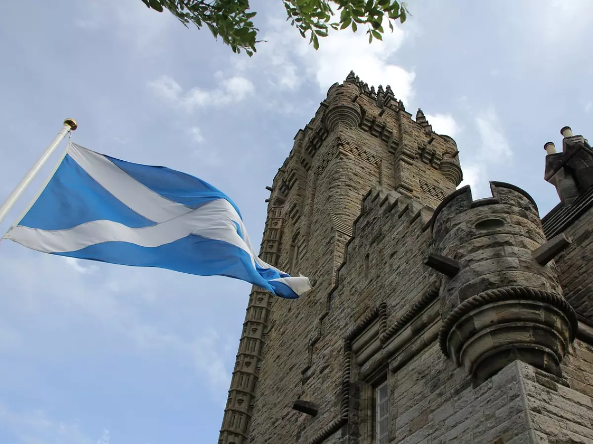 A tall, stone tower with a pointed spire, with the Scottish flag flying in the wind in the foreground. 