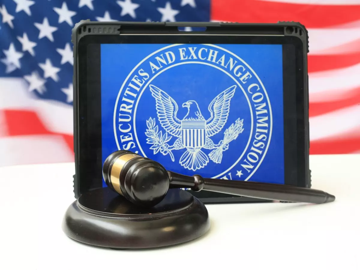 A close-up of a tablet displaying the Securities and Exchange Commission (SEC) logo, accompanied by a wooden gavel and a soundboard. A blurred American flag serves as the backdrop.