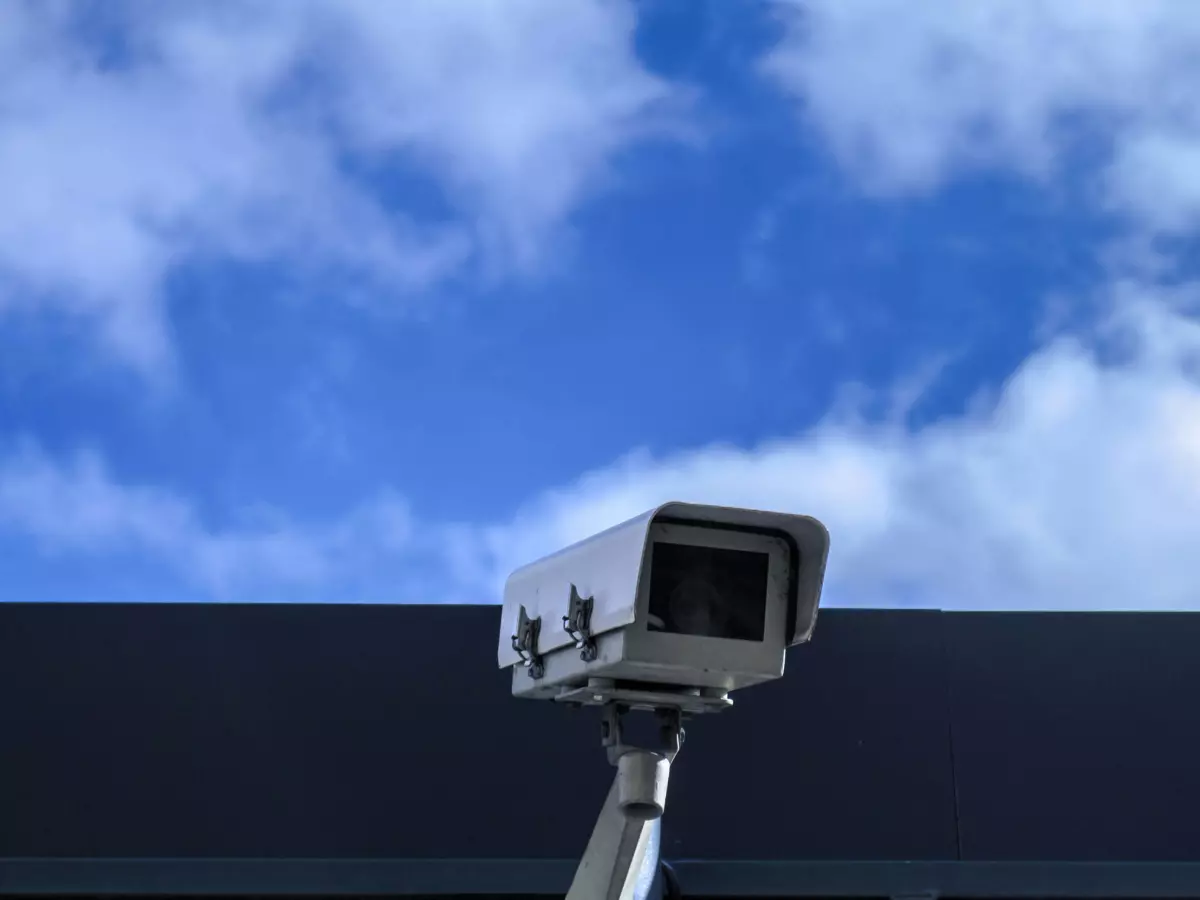 A security camera mounted on a building with a blue sky and white clouds in the background.