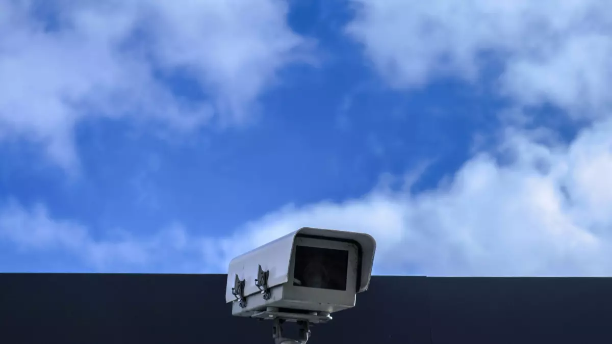 A security camera mounted on a building with a blue sky and white clouds in the background.