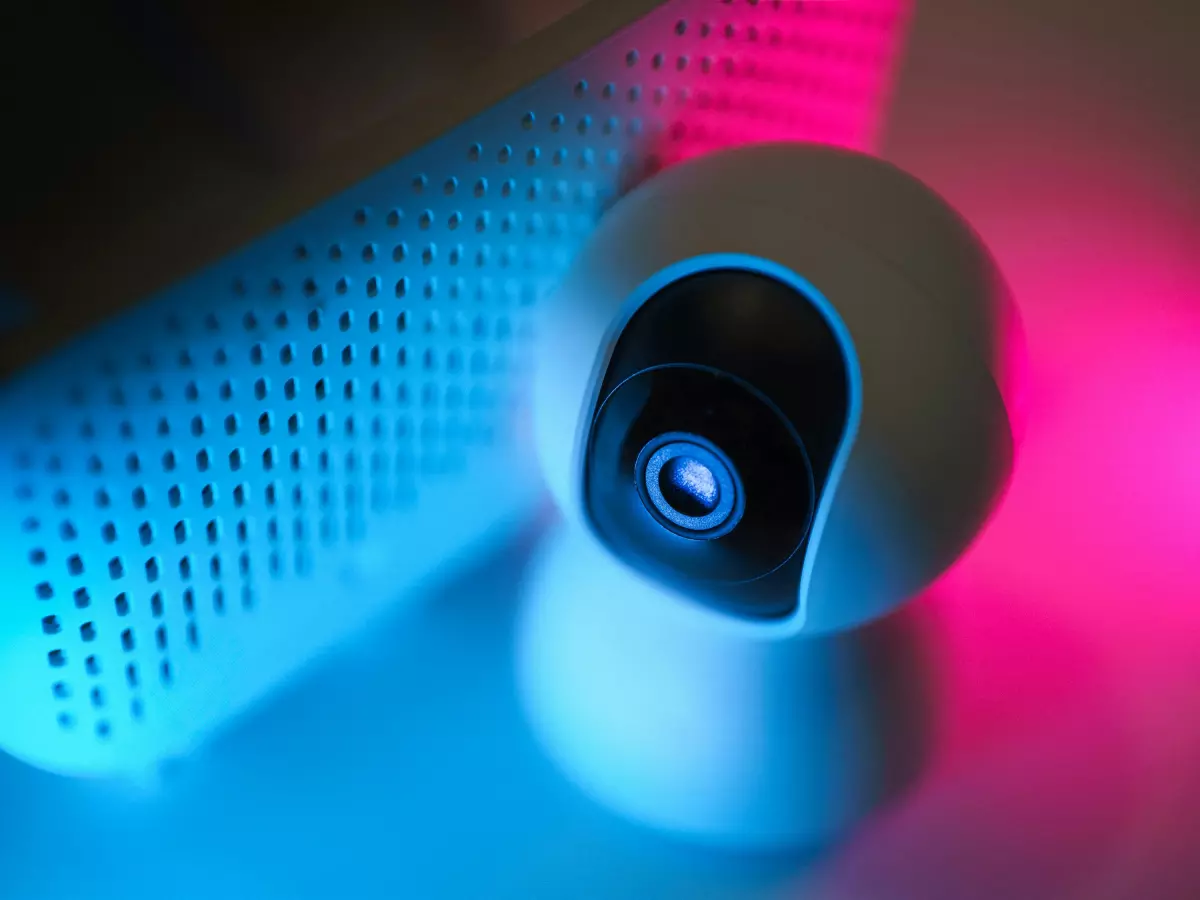 A close-up shot of a white security camera with a black lens. The camera is illuminated by blue and pink light.