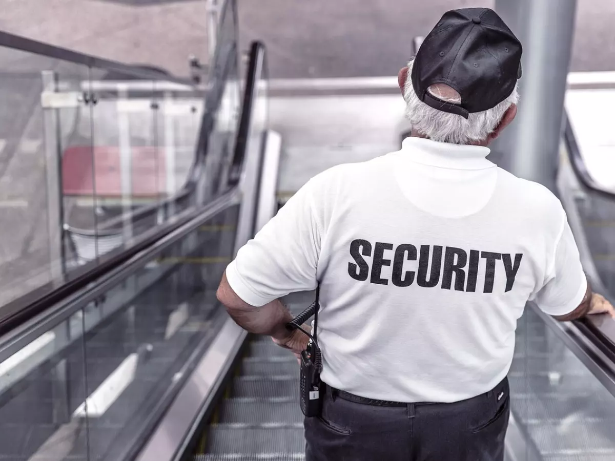 A security guard in a white shirt with "SECURITY" written on the back, walking down an escalator.  The guard is looking forward and his face is not visible.