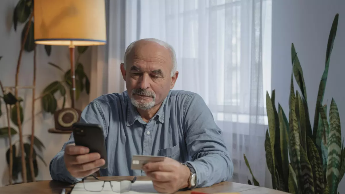 A person holding a smartphone and looking at it with a neutral expression. The person is sitting at a table with a glass of water and a notebook in front of them.