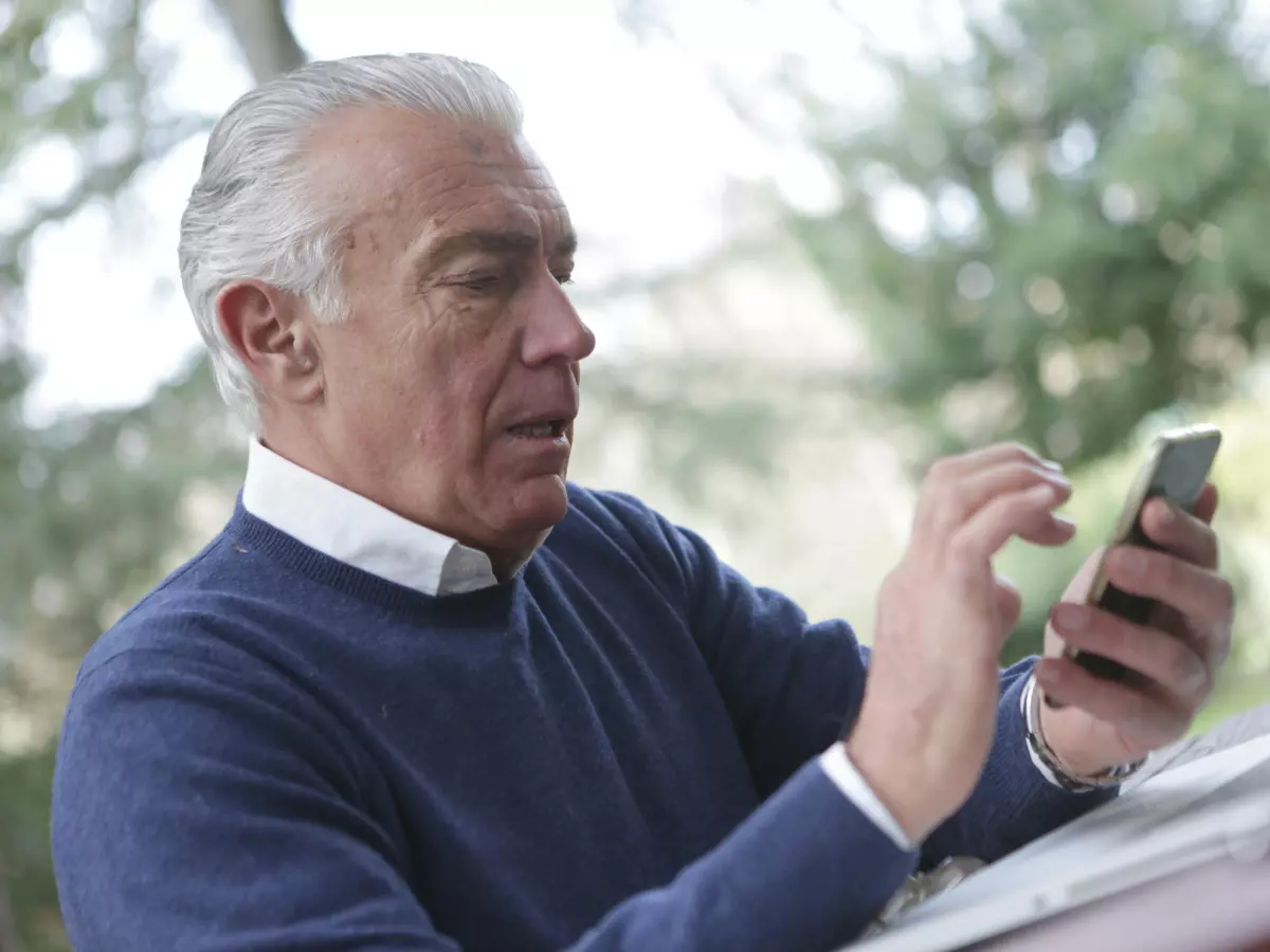 A man is sitting at a table with a laptop in front of him. He is looking at his phone. He is using a smartphone.  