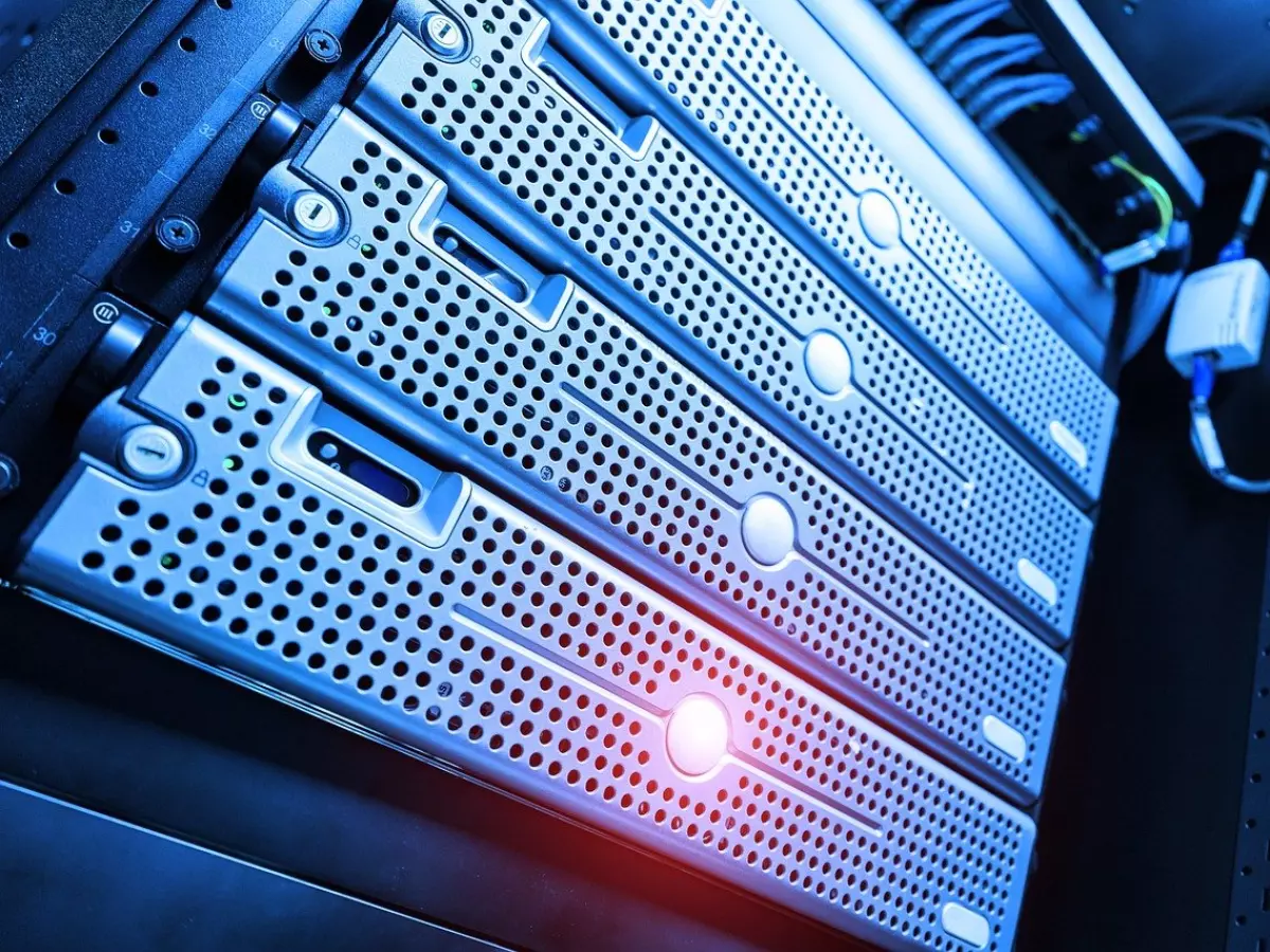 A close-up shot of a server rack with multiple server units stacked on top of each other. The servers are silver and have a textured finish with a pattern of small holes. The front of the servers is visible, showing the lights and buttons. The background is blurred, and there is a network cable and a device attached to the servers.