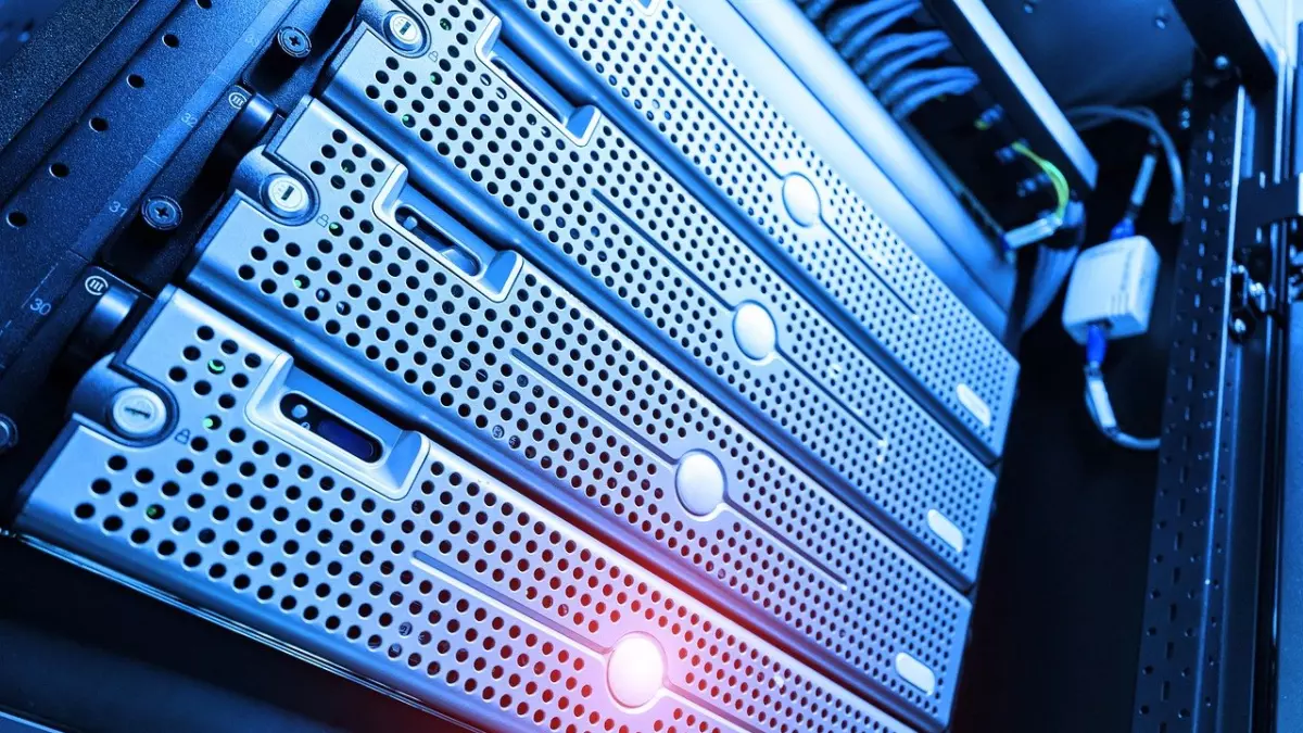 A close-up shot of a server rack with multiple server units stacked on top of each other. The servers are silver and have a textured finish with a pattern of small holes. The front of the servers is visible, showing the lights and buttons. The background is blurred, and there is a network cable and a device attached to the servers.