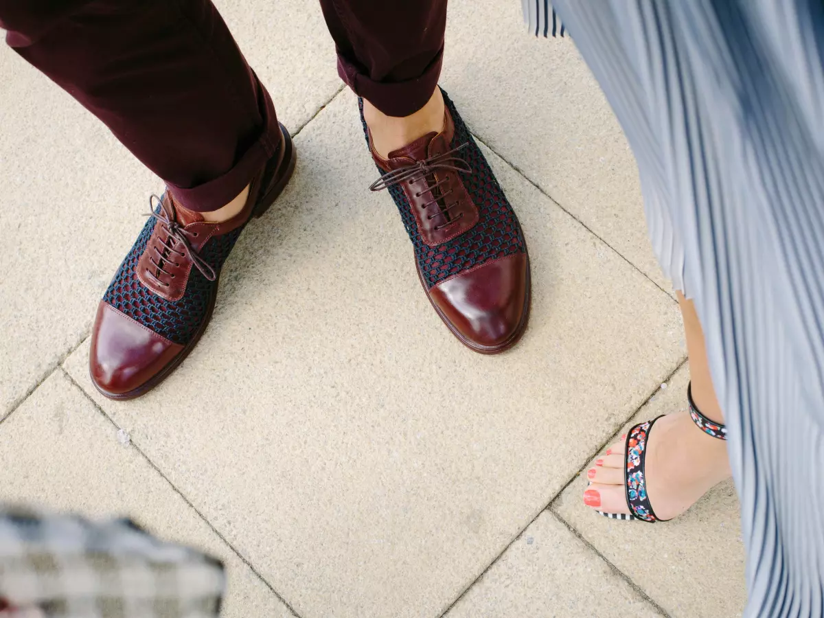 A pair of brown leather shoes with white laces are worn by a man. The other shoe is visible.  A pair of women's feet in sandals are also visible in the bottom right corner.