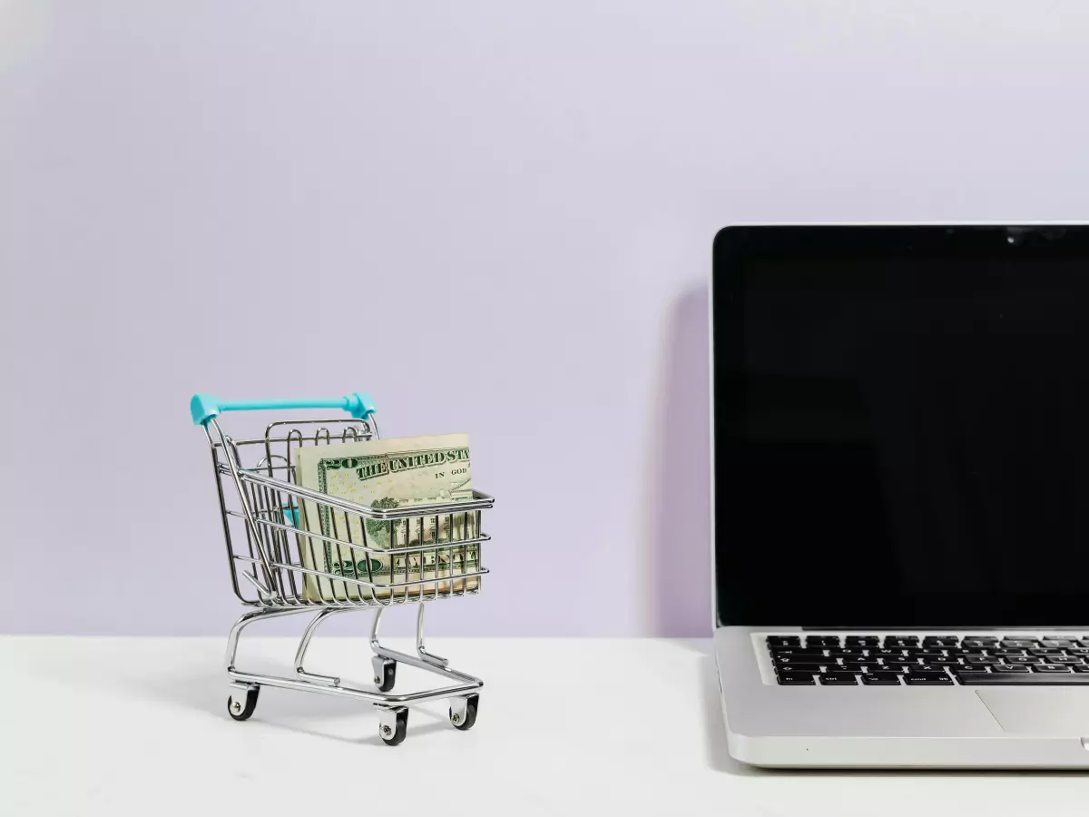 A miniature shopping cart with a stack of dollar bills inside, placed next to a closed laptop on a white surface against a light purple wall.