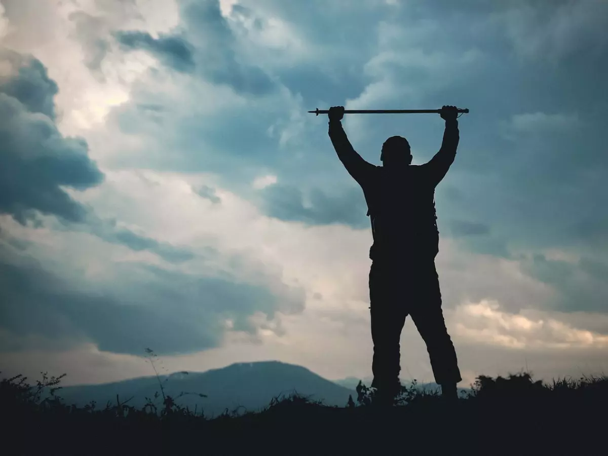 A man in silhouette stands on a mountaintop, holding a staff high above his head. The sky is dramatic and cloudy.