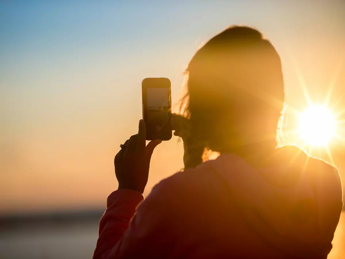 A person silhouetted against the sunset is taking a photo with a phone.