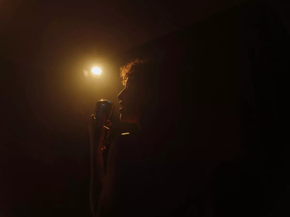 A person speaking into a microphone, with only their face and the microphone illuminated in a dark room.