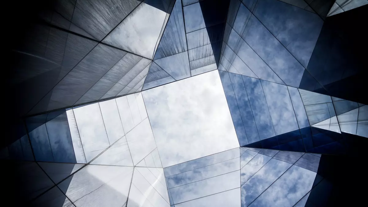 A view of the sky through the glass panels of a modern building. The building is made of glass and steel, and it is surrounded by a cloudy sky. The image is taken from a low angle, so the building appears to be very tall. The image is in color and the focus is on the sky.