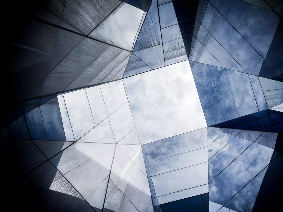 A view of the sky through the glass panels of a modern building. The building is made of glass and steel, and it is surrounded by a cloudy sky. The image is taken from a low angle, so the building appears to be very tall. The image is in color and the focus is on the sky.