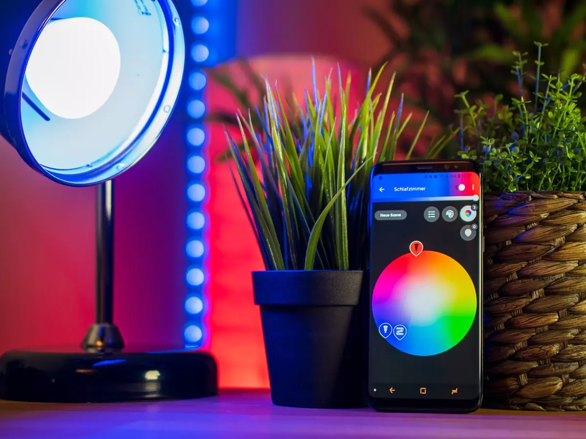 A smartphone is shown in the foreground, with a smart home control app open on the screen. In the background, a smart light bulb, a strip of smart LED lights, and two potted plants are visible. The smartphone's screen is displaying a color picker wheel, which can be used to adjust the color of the smart lights.