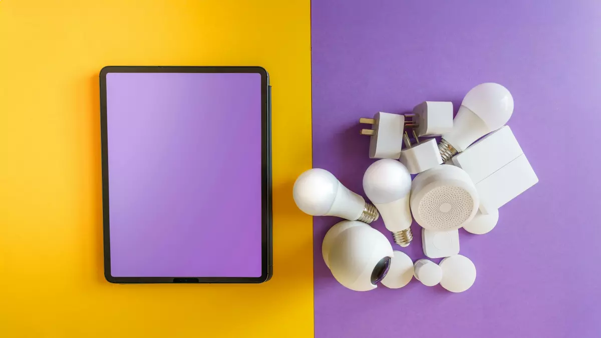 A flat lay of a tablet and a variety of smart home devices, including smart bulbs, smart plugs, and a smart thermostat, on a yellow and purple background.