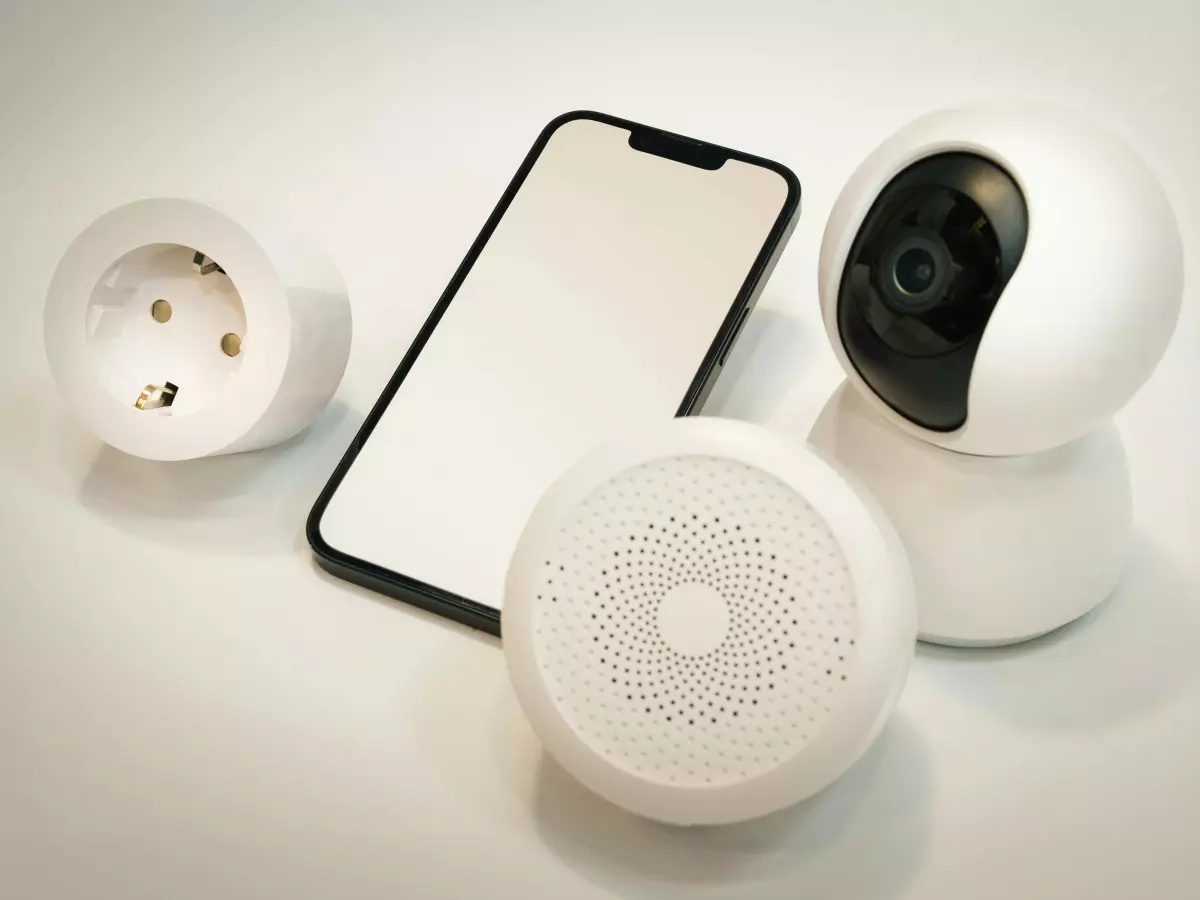 A smartphone lies at the center of the image, flanked by a smart security camera, a smart plug, and a smart speaker. All devices are white, creating a cohesive visual.