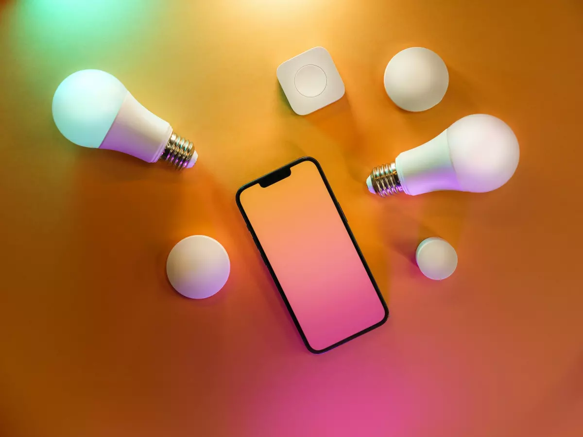 A close-up shot of a smartphone and several smart home devices including lightbulbs and a smart home hub, set against a colorful background.