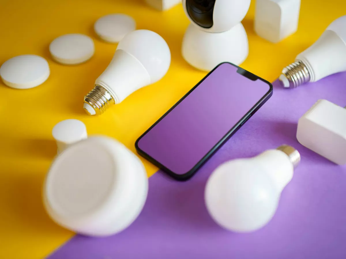 A smartphone lies on a split yellow and purple background surrounded by Philips Hue light bulbs and accessories.  The bulbs are white and the smartphone is black.