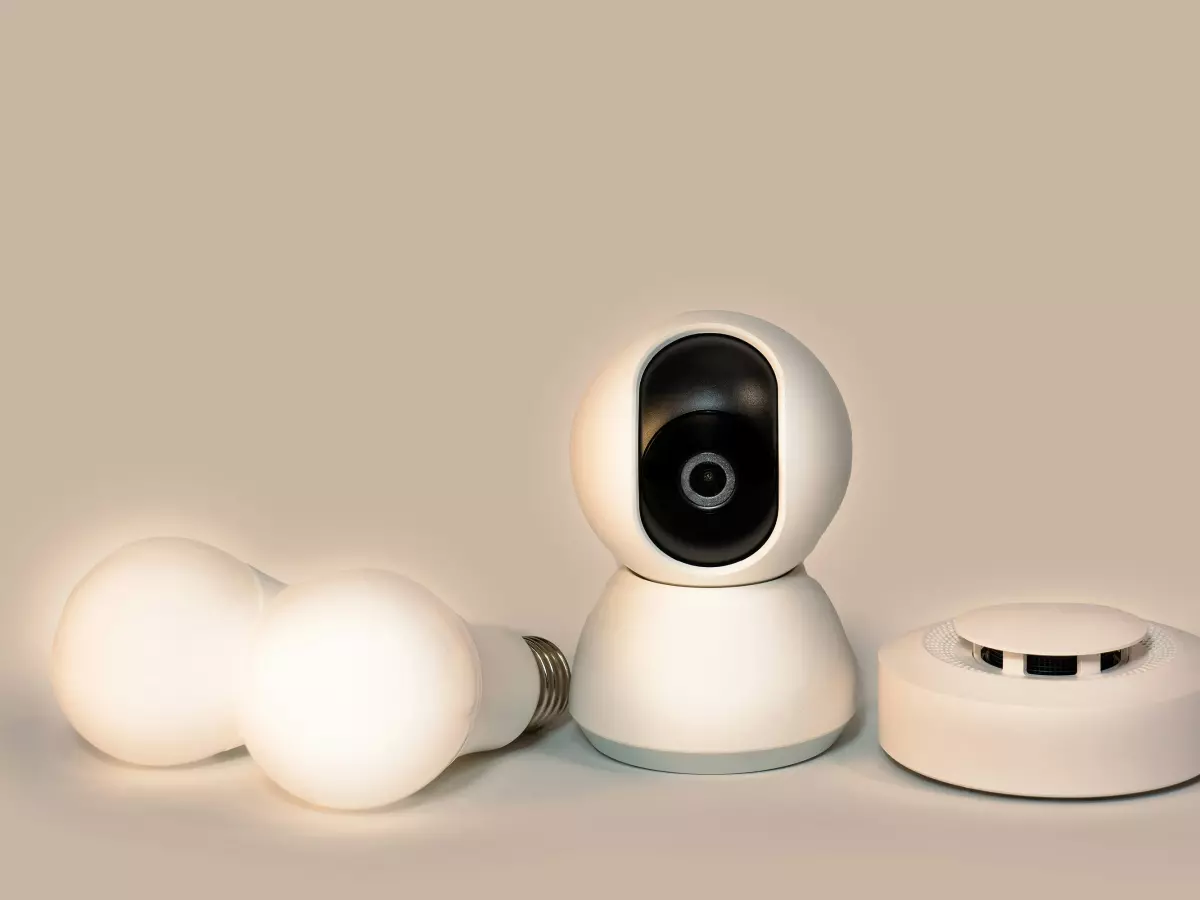 A security camera, two smart bulbs, and a smoke detector all in white, arranged in a row on a light brown background.