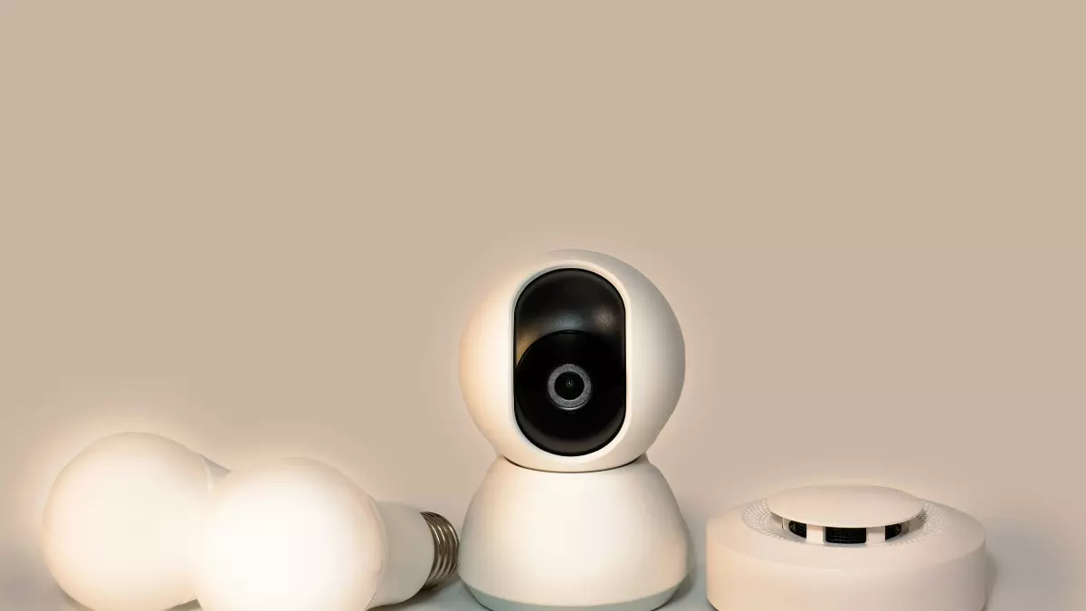 A security camera, two smart bulbs, and a smoke detector all in white, arranged in a row on a light brown background.