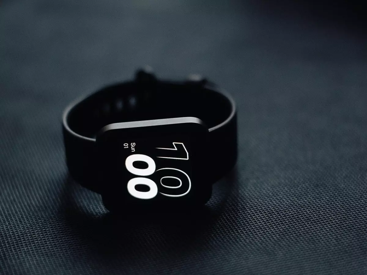 A black smartwatch on a dark textured surface. The screen displays the time and date.