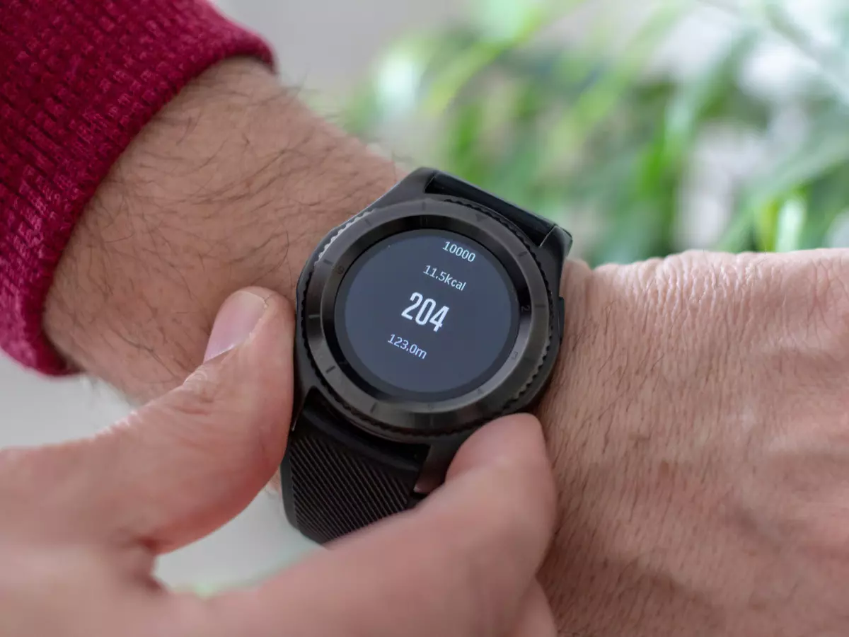 A man's hand is shown pressing the screen of a smartwatch. The watch is displaying various readings including heart rate.