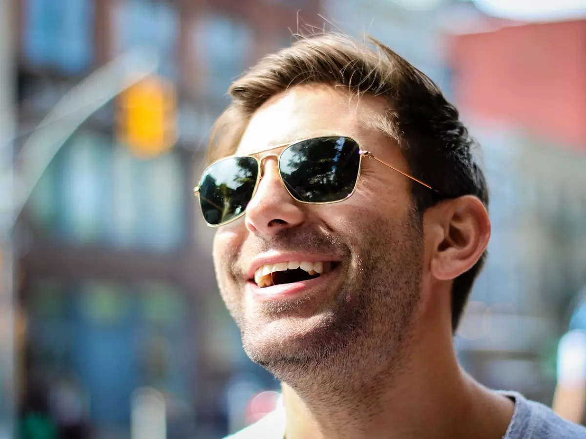 A man wearing sunglasses and a blue shirt looks up and smiles.