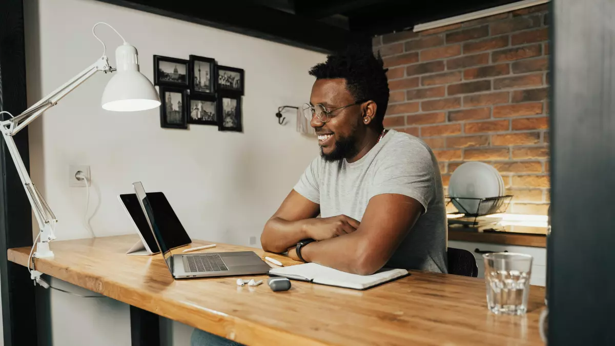 A man sits at a desk with a laptop and a notebook. He is smiling and looking at something off-screen.