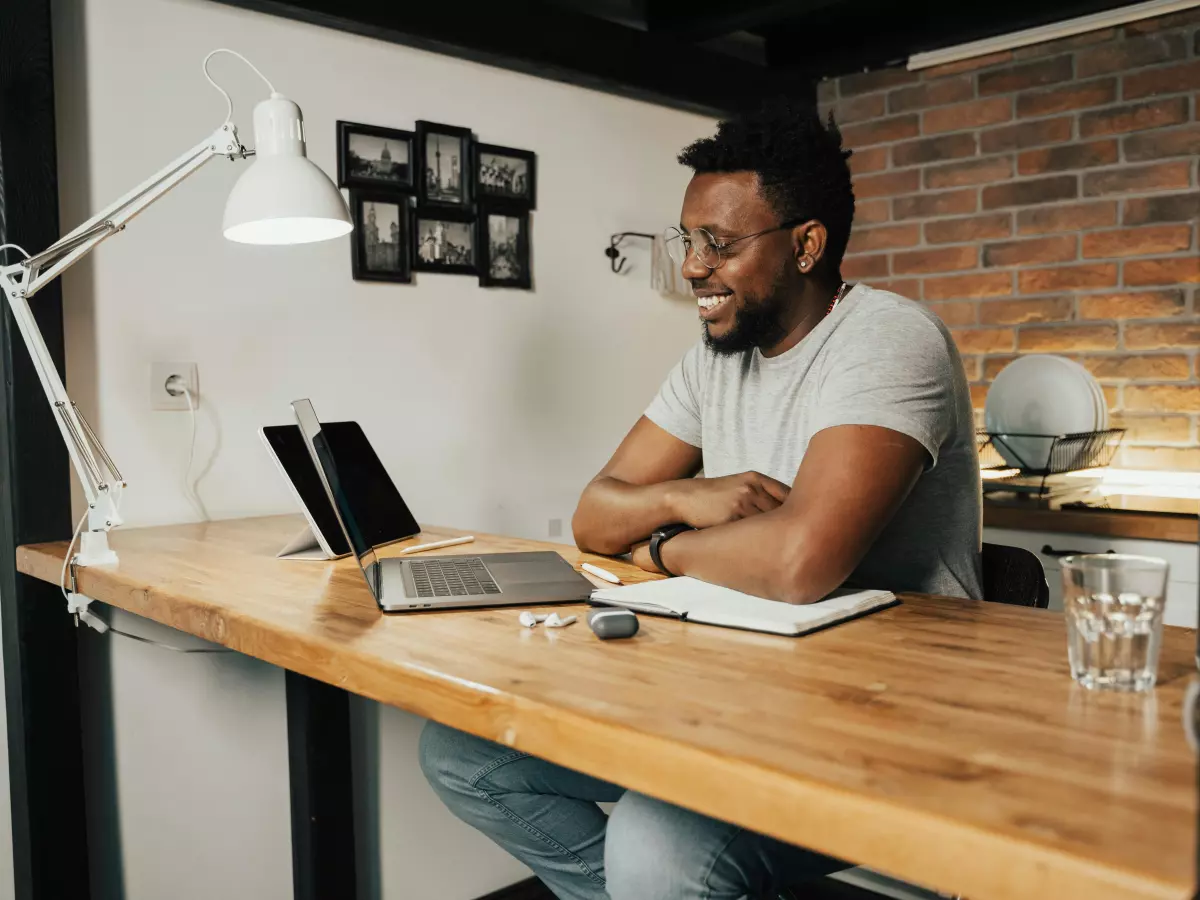 A man sits at a desk with a laptop and a notebook. He is smiling and looking at something off-screen.